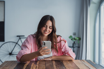 Smiling businesswoman looking at phone screen, sitting at work desk, positive young woman chatting...