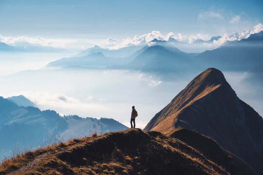 Woman with backpack standing on a mountain ridge above clouds in the Swiss Alps at sunset. Scenic view of alpine peaks, silhouette of girl, fog, clear sky in fall. Hiking, travel, outdoor adventure