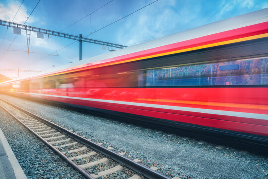 Blurred red passenger train passing mountain railway station in Swiss Alps at sunset. Moving high-speed train. St. Moritz, Switzerland. Bernina Express. Railway platform, cloudy sky. Rail transport
