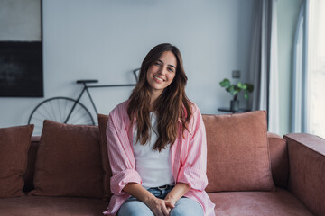 A happy young woman sits on a sofa in her modern cozy home, smiling at the camera, relaxing alone,...