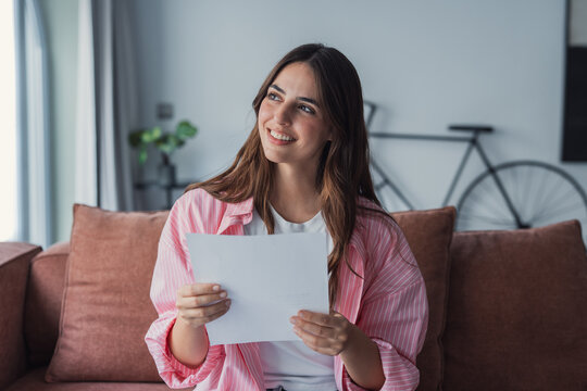 Relieved young woman sitting on couch reading medical test results at home. Smiling female holding healthcare document with good news from doctor or clinic.