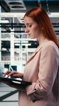 Vertical video Optimizing electronics, laptop and woman in server farm doing improvements. Monitoring system performance, notebook and female technician walking in data center, identifying bottlenecks