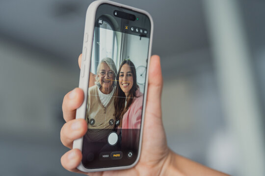 A happy young woman and her mature mother take a selfie together on a smartphone, smiling and having fun, with the phone screen visible showing their joyful expressions while capturing the moment 