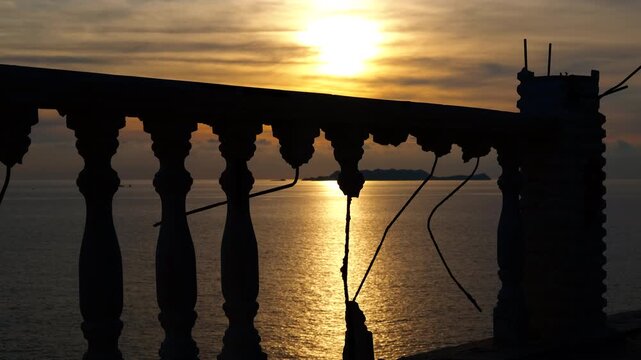 Golden sunrise reflecting on the sea from a balcony. Beautiful golden sun rising over the calm ocean, viewed through the silhouette of a stone balustrade on an abandoned balcony