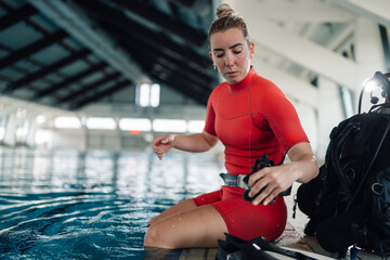 Female scuba diver preparing for training, sitting by the indoor pool edge, wearing a red wetsuit...