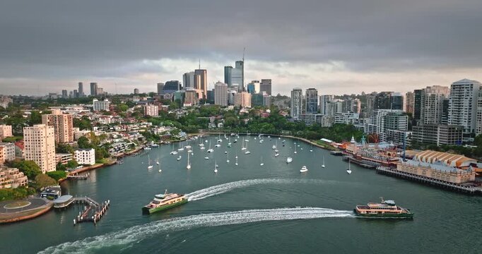 Sydney cityscape and North Sydney suburbs flanking Lavender Bay, calm inlet filled with moored yachts, two ferries navigate the water, creating wakes under a cloudy sky. Aerial drone zoom out panorama