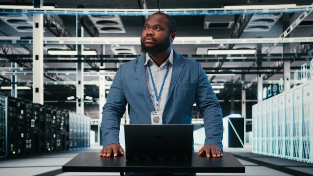 Server farm programmer at workplace desk using laptop to optimize electronics. African american man in data center using notebook to monitor system performance, identifying bottlenecks, camera A