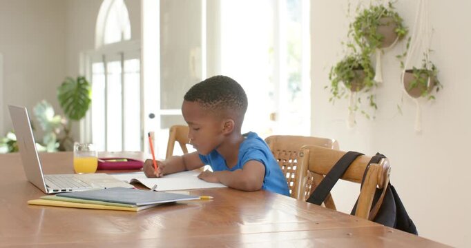African American child studying with red pencil at dining table as parent coming adjusting backpack