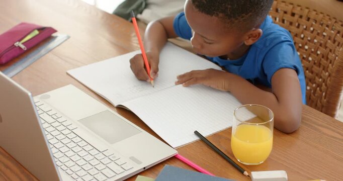 Child is writing homework with red pencil at wooden table, adult entering and placing mug nearby
