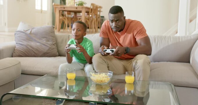 African American father and son playing game on sofa after game start, controllers, chips, juice