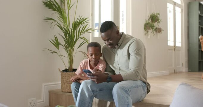 African American father and son sitting on low step at home, father tapping smartphone, guiding son