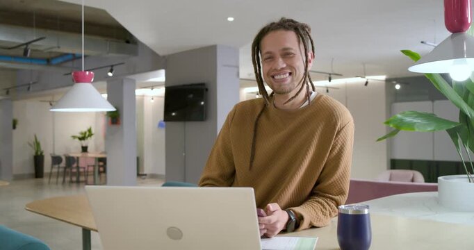 African American man laughing and smiling after camera gaze while holding phone by laptop in lounge