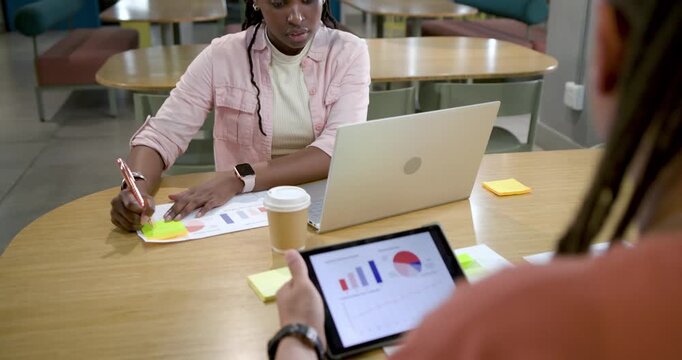 African American pair annotating charts at table, man shifting tablet toward woman for data review