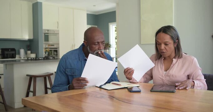 Mature couple sitting at wood table comparing papers and tablet, reviewing documents, woman tapping