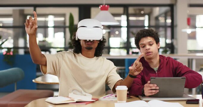 African American men at study table one donning white VR headset gesturing as friend guiding tablet
