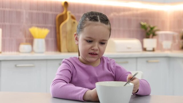 Unhappy little girl refusing to eat porridge at light table in kitchen