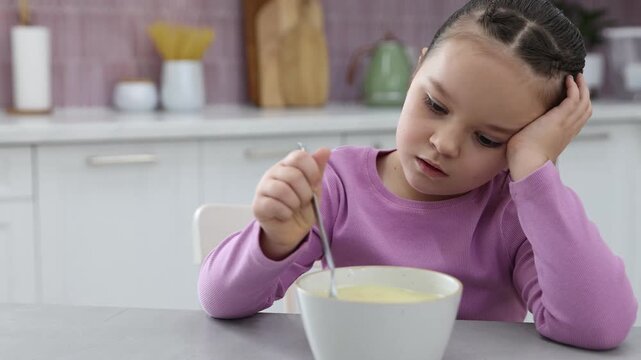 Unhappy little girl refusing to eat porridge at light table in kitchen. Camera moving forward