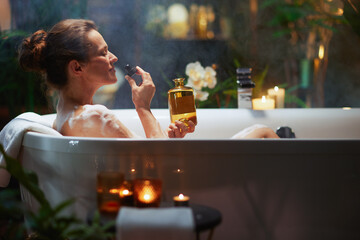 A woman enjoys a luxurious bubble bath, eyes closed in serenity, as she smells an essential oil from a dropper bottle. Candles, plants, and warm light create a tranquil, pampering home spa experience. © Alliance