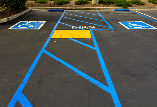Accessible parking spaces with blue lines and symbols flank a striped no parking zone with a yellow ramp on a clean asphalt lot bordered by greenery and curbs