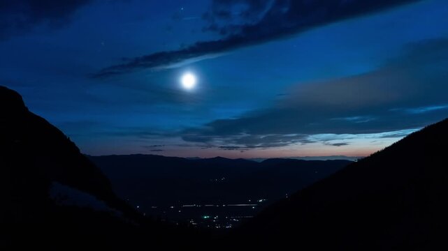 Bright moon setting through clouds in a blue twilight sky above a mountain valley with town lights, Pine Creek Lake, Montana, time lapse