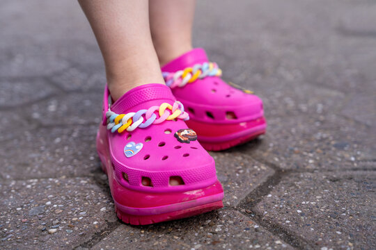 Belgrade, Serbia: July 3, 2024: Close up of legs wearing bright pink rubber clogs crocs decorated with multicolor chain and small shoe charms, standing on textured pavement outdoor daylight in summer
