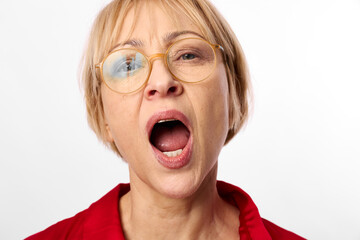 Middle aged woman with short blonde hair and glasses yawns widely wearing a red shirt. Close-up on white background. Concept of tiredness, fatigue, and everyday life emotions.