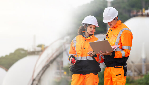 Technical team in safety uniform discussing maintenance plan at fuel storage facility, Industrial engineers using laptop and radio to monitor gas storage tank system