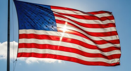 Fototapeta premium A large American flag waves proudly against a bright blue sky with scattered white clouds. The sun shines brightly behind the flag, creating a radiant effect.
