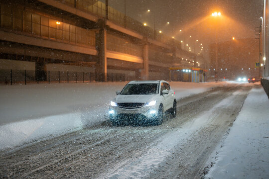 Heavy snowfall and blizzard in city, traffic roads covered with thick layer of snow and ice, car driving through snowy frosted roadways in evening under street lights, winter weather conditions.