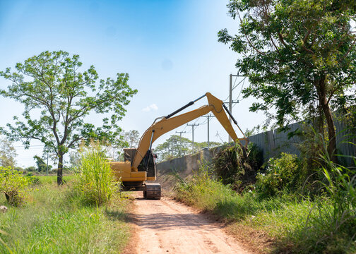 A yellow excavator performing land clearing and site preparation work along a rural dirt road, removing overgrown vegetation near a metal fence on a sunny day.