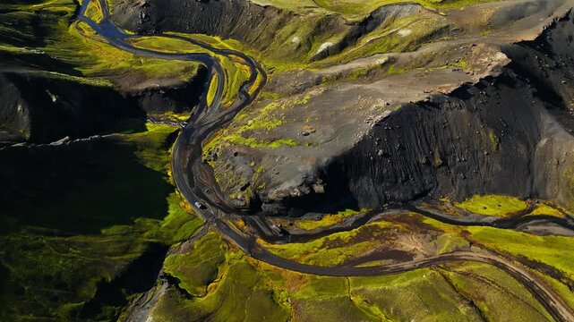Aerial drone shot of a tiny car driving a winding mountain road through green Nordic highlands and dark volcanic cliffs. Epic summer road trip scenery and remote travel escape.