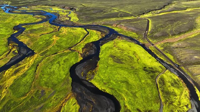 Drone aerial of a tiny car driving a remote road beside a winding river through bright green Icelandic highlands. Epic summer journey, solitude, and wild nature travel concept.