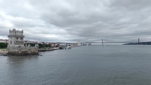 Belem Tower on Tagus river bank with Ponte 25 de Abril suspension bridge in background under rainy sky, Lisbon, Aerial, Wide shot