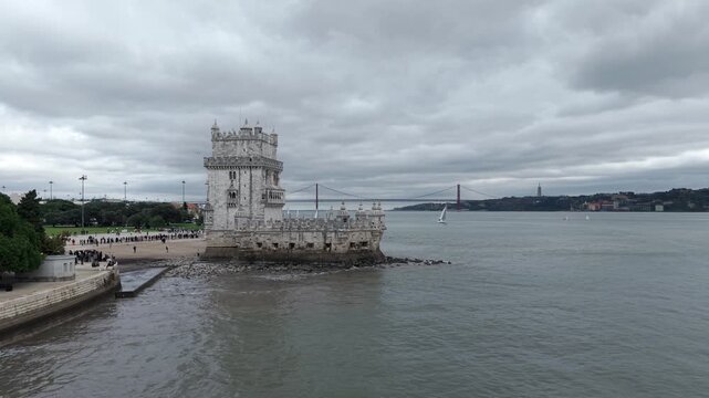 Belem Tower on Tagus river bank with Ponte 25 de Abril suspension bridge in background under rainy sky, Lisbon, Drone shot