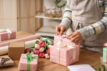 Young woman with tulip flowers and packing materials tying bow on wrapped gift boxes for...