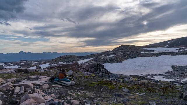 Backpacker resting at an alpine camp among rocks and snowfields as clouds move through a golden sunset, Beartooth Plateau, Montana, time lapse
