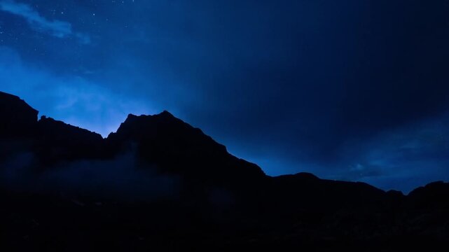 Lightning flashes lighting up clouds behind a dark mountain silhouette under stars at night, Beartooth Mountains, Montana, time lapse