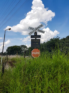 A Portuguese railway crossing sign with a crossbuck reading RAILWAY CROSSING above a stop sign STOP, photographed in a rural landscape under a bright blue sky with clouds.