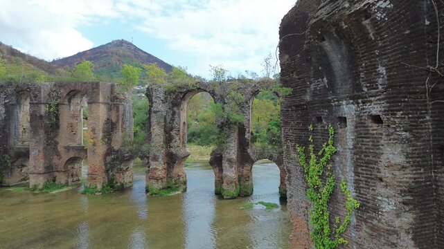 Cinematic drone slide reveal from an ancient brick wall to the monumental arches of the Roman Aqueduct of Nicopolis in Epirus Greece featuring historic masonry and scenic river landscape.
