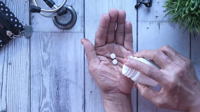Unrecognizable senior woman taking multiple pills from bottle on her palm on rustic wooden table with stethoscope, plant, and spectacles