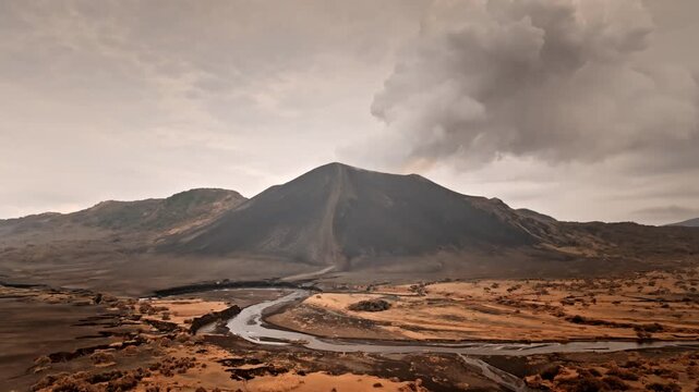 Mountain Yasur volcano is actively erupting, sending large ash cloud into the sky above the barren volcanic landscape, with a river flowing through the wide valley and dry brush. Aerial drone flight