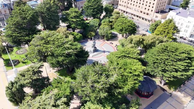 Aerial circling Plaza Mu&ntilde;oz Gamero in Punta Arenas, Chile, featuring the Magellan Monument surrounded by lush cypress trees and historic city architecture
