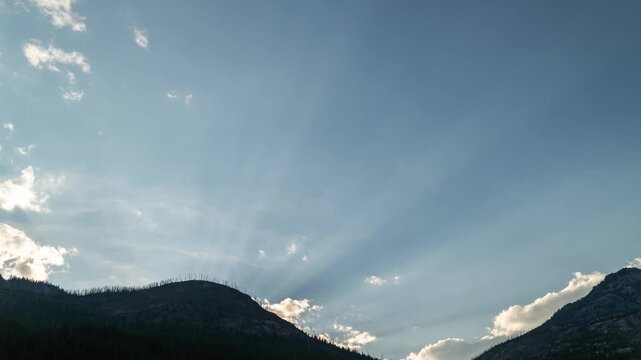 Sun rays streaming from behind a silhouetted mountain ridge into a blue sky with drifting clouds, Montana, time lapse