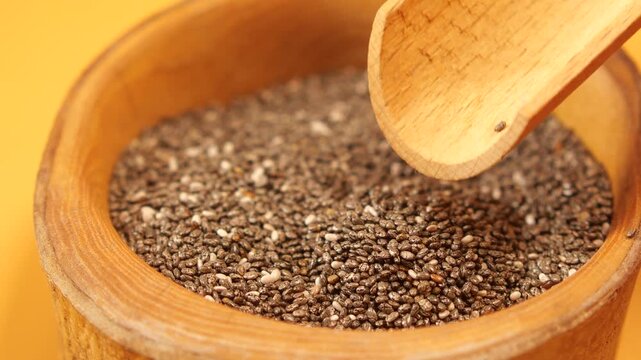 Chia seeds pouring from wooden scoop into round bamboo bowl on yellow background
