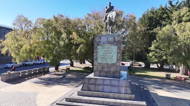 Aerial rises then circling the equestrian statue of General Manuel Bulnes in Punta Arenas, Chile, featuring the stone pedestal and lush greenery of Avenida Bulnes
