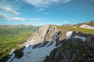 Scenic aerial dizzying top view to rocky cliff and sharp rocks under clouds in blue sky. Vertigo vivid alpine scenery with sheer crags above small lake in green dale. Colorful dizzy high mountains. © Daniil