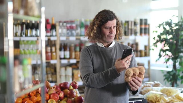 Customer in a European store holds a package of eggs in his hands and scans a barcode on a mobile phone. Man uses an online application to pay by QR code. High quality 4k footage