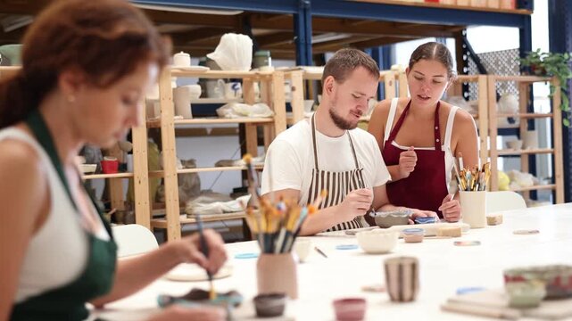 Female teacher and mentor helps students learn how to make patterns on ceramic plates using paint and brush in a pottery workshop. High quality 4k footage