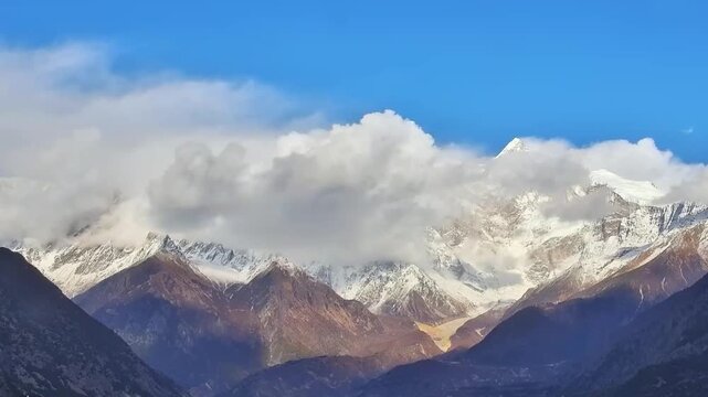 A breathtaking aerial timelapse capturing the daylight transitioning into a golden sunset over the snow-capped peak of Mount Namcha Barwa, with clouds swirling around the summit in Tibet, China.
