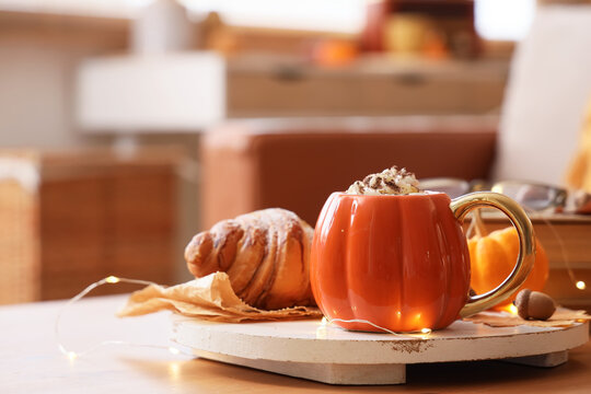 Cup of pumpkin latte with croissant and glowing lights on table in room, closeup